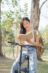 Young asian women with backpack sitting on bicycle and reading a book with happiness to resting after cycling bicycle in the nature park while relaxation with journey travel for healthy lifestyle