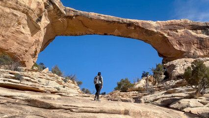 Female hiker walking up a rocky trail towards a natural sandstone formation known as Owachomo Bridge with a bright blue sky behind. Natural Bridges National Monument, Utah, USA.