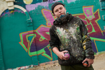 Young man in black stylish hoodie looking at camera while standing against green brick wall with graffiti artwork