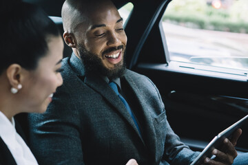 Cheerful African American male director communicating with female secretary during car drive to business meeting discussing startup ideas and reading notes for presentation from digital tablet