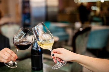 Two hands holding wine glasses, one with red and the other with white wine, toasting in a blurred restaurant background. Cheers!