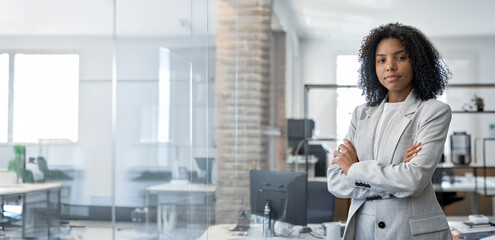 Young African American woman business leader manager, standing confident with crossed arms, looking at camera in business office. Portrait of professional businesswoman in suit. Banner, copy space