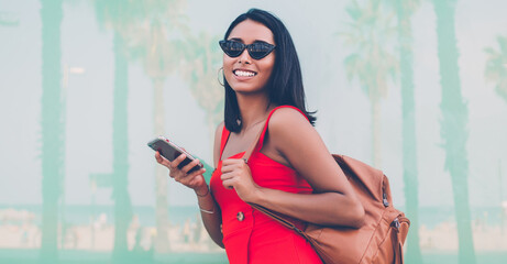 Obraz premium Half length portrait of cheerful woman in fashionable sunglasses holding mobile phone for chatting in hands and laughing at camera during day for walking, gladden jubilate hipster girl phoning
