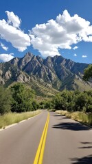 Naklejka premium A vast yellow road leads towards majestic mountains, illuminated by sunlight, flanked by lush greenery and a stunning blue sky with scattered clouds overhead