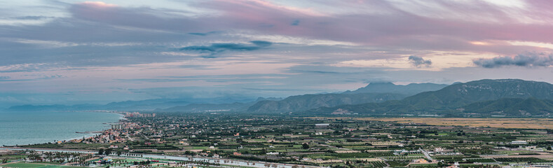 Obraz premium Wide panoramic view of a coastal town with buildings, green fields, and mountains stretching across the horizon near the sea.