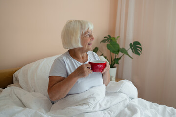 Relaxed Caucasian senior blonde hair woman in white pajama sitting on bed and drinking latte coffee...