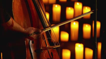 Close-up of a musician playing a bow on a stringed instrument. Candles burning in the background. The light of candles creates a serene and intimate atmosphere of the concert.