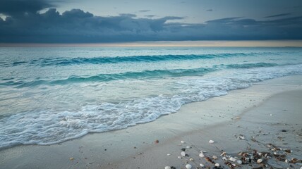 A photograph of a beautiful soft blue ocean wave gently lapping against the fine sandy beach. The water is clear, and the sand is lightly dotted with seashells. The sky is overcast, with dark clouds