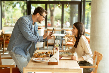 Couple enjoying a cozy meal together in a bright cafe, capturing memories with a smartphone during a sunny afternoon