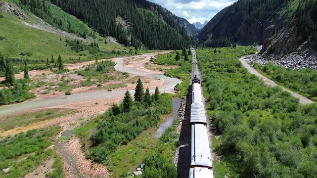 Close up flyover drone shot of scenic passenger train departing Silverton, CO, and approaching a canyon. Flight shows a river in the foreground with mountains in the background.