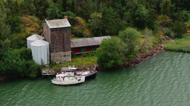 Two boats glide toward a weathered grain elevator along the Aland islands' coast in Finland. Lush green trees and rocky shores frame this picturesque landscape