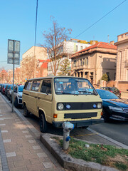 Obraz premium Vintage van car is parked along a bustling city street, surrounded by urban architecture and clear blue skies. Scene captures a blend of nostalgia and everyday life in a lively neighborhood