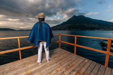 Indigenous man with Andean hat and blue poncho standing on a wooden dock, looking at a lake during sunset under a cloudy sky with mountains in the background.