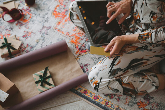 Overhead view of a young woman sitting on the floor in pyjamas surrounded by wrapped gifts and wrapping paper holding a digital tablet ordering gifts