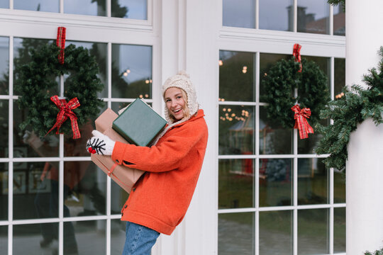 Smiling woman carrying a stack of wrapped Christmas gifts standing outside a house decorated for the holidays