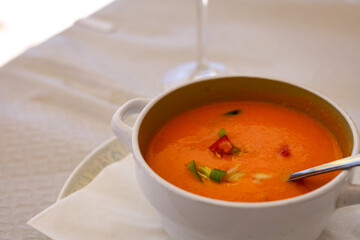 Horizontal image of a white bowl filled with traditional gazpacho, a cold Spanish tomato soup, topped with diced tomatoes, greens. Smooth texture, vibrant colors create a fresh culinary presentation