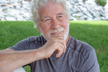 Portrait of attractive smiling senior man of 70 years old sitting in the park looking at side. Elderly grandfather with white hair and beard relaxing enjoying a sunny day in outdoors