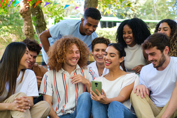Group of diverse young adults are watching funny content on mobile phone at summer festive party outdoors and laughing