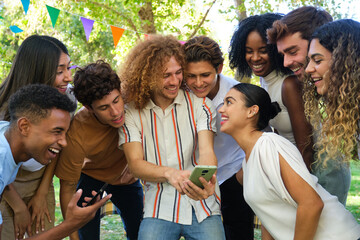 Happy friends laughing and having fun using a smartphone outdoors in a park