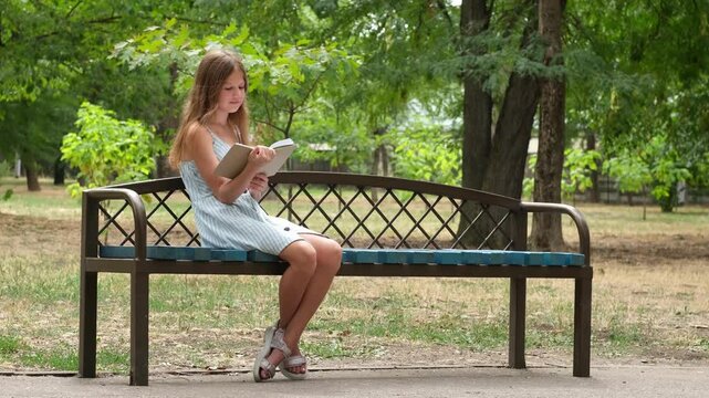 Cute smiling preteen girl reading book on bench in city park, resting outdoors. Green nature background