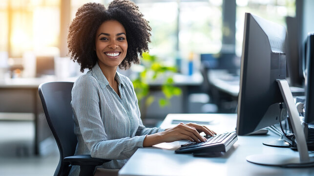 Smiling businesswoman sitting in front of a computer in a modern office, surrounded by greenery, creating a pleasant and productive work atmosphere.