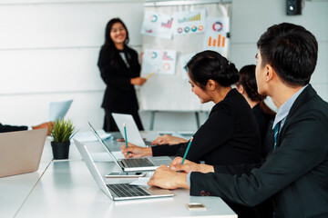 Asian business people listening to presentation from businesswoman who stands in front of group meeting room at international business conference. Multicultural corporate strategy concept. uds