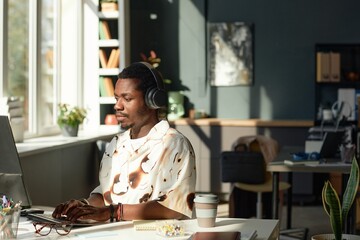 Wide angle side view of young Black man using computer in sunlight and wearing big noise cancelling headphones in office interior, copy space 