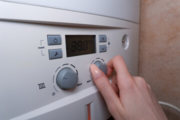 A close-up of a woman's hand with a manicure, turning the knob on the gas boiler to regulate the water temperature in the house. The control panel is clearly visible, showing precise settings for hot 