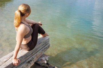 Back view of young woman in sportswear sits on a wooden beam on the shore of a lake