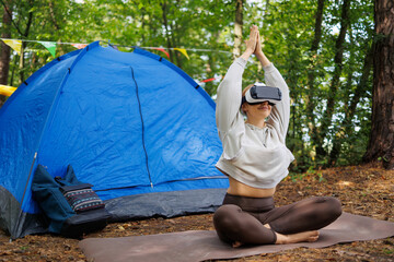 Young athletic woman in hoodie and leggings sits meditating on yoga mat against background of tent in forest