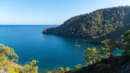 Naklejka premium A yacht in the stunning Turkish Mediterranean coastline in Antalya, inside a blue sea, surrounded by lush green forests, and rugged rocky shores, attracting tourists every summer.