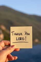 Hand holding handwritten note: Thank You LORD with river water and mountain in the background....