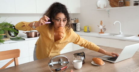 Hurrying young African-American woman with croissant and laptop applying mascara in kitchen