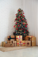 Decorated Christmas tree with balls, glowing lights and gift boxes near white brick wall