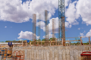 Construction site under a clear blue sky with clouds. Several tall, vertical steel rebar structures are in place for future concrete columns. In the foreground, concrete foundation.