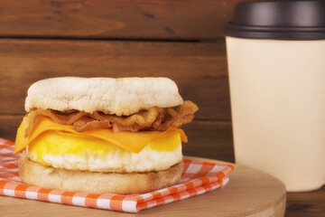 Sandwich breakfast, cup of coffee and wooden plank in background. English muffin, egg, cheese, and bacon.