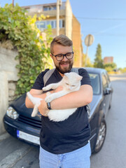 Bearded man gently embraces a white cat, conveying a moment of warmth and connection with pet. Outdoor setting adds an element of everyday life and companionship