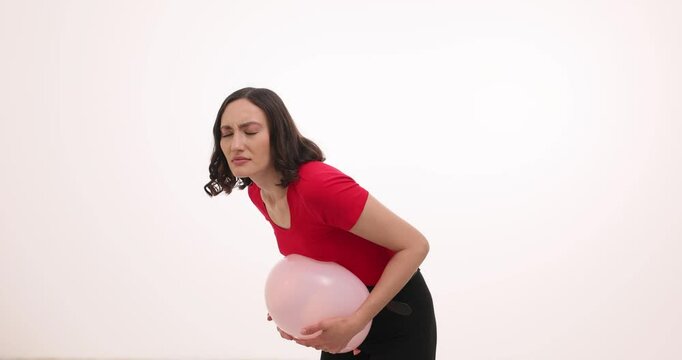 Young woman in red holds balloon near belly experiencing discomfort on white background. Lady shows metaphor of digestive problems and period pain