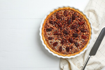 Delicious pecan pie in baking dish and knife on white wooden table, flat lay. Space for text