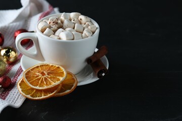 Tasty hot cocoa drink with marshmallows in cup, dry orange slices and baubles on black table. Space for text