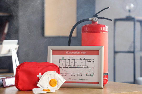 Evacuation plan with masks, first aid kit and fire extinguisher on table in burning office building