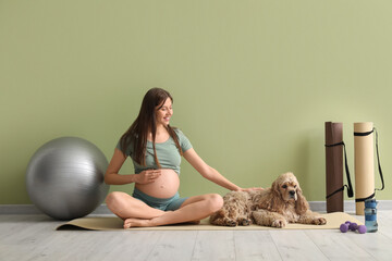 Beautiful young pregnant woman with cocker spaniel and sports equipment practicing yoga near green...