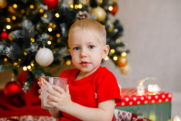 A toddler boy in a red sleeveless shirt enjoys milk and cookies beneath the Christmas tree