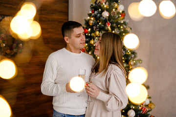 A couple in love hugs and sips champagne in front of Christmas tree with bokeh from twinkling...