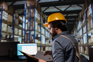 Depot worker examining products orders on laptop and reading dispatch notes before approving stock...