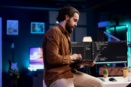 IT engineer doing software debugging using laptop, checking glitches in lines of code displayed on desktop PC monitors. Man standing on desk at home fixing coding issues using notebook