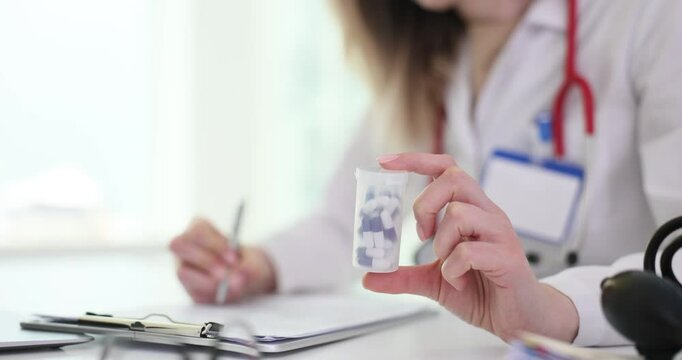 Doctor holds medicine bottle writing prescription on clipboard in office setting. Blonde physician recommends treatment at healthcare facility