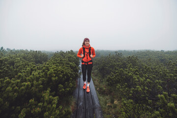 A smiling woman in an orange puffy jacket stands by a misty lake surrounded by greenery. She wears a headband and has a relaxed posture, enjoying the serene outdoor environment. The fog creates a tran