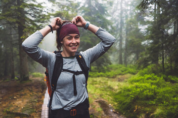 A young woman with a headband smiles while adjusting her hair in a misty forest. She wears a long-sleeve shirt and a backpack, surrounded by lush greenery and trees. The atmosphere is serene and adven