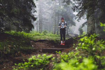 A woman in an orange jacket and red sneakers sits on a wooden bench in a foggy forest. She appears relaxed, surrounded by lush green grass and trees. The atmosphere is serene and tranquil, perfect for